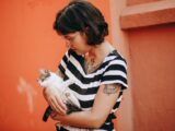 Woman with tattoos holds a kitten in striped dress against orange backdrop.