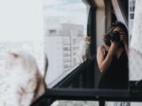 Woman photographing cats on a city balcony with a blurred cityscape in the background.